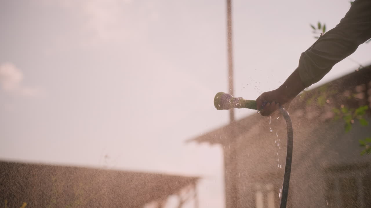 손으로 물 프를 들고 공중으로 안개를 뿌리는 클로즈업, 빛이 물방울에 부드러운 빛을 만들어, 배경에는 흐릿한 건물과 초록색이 있습니다.