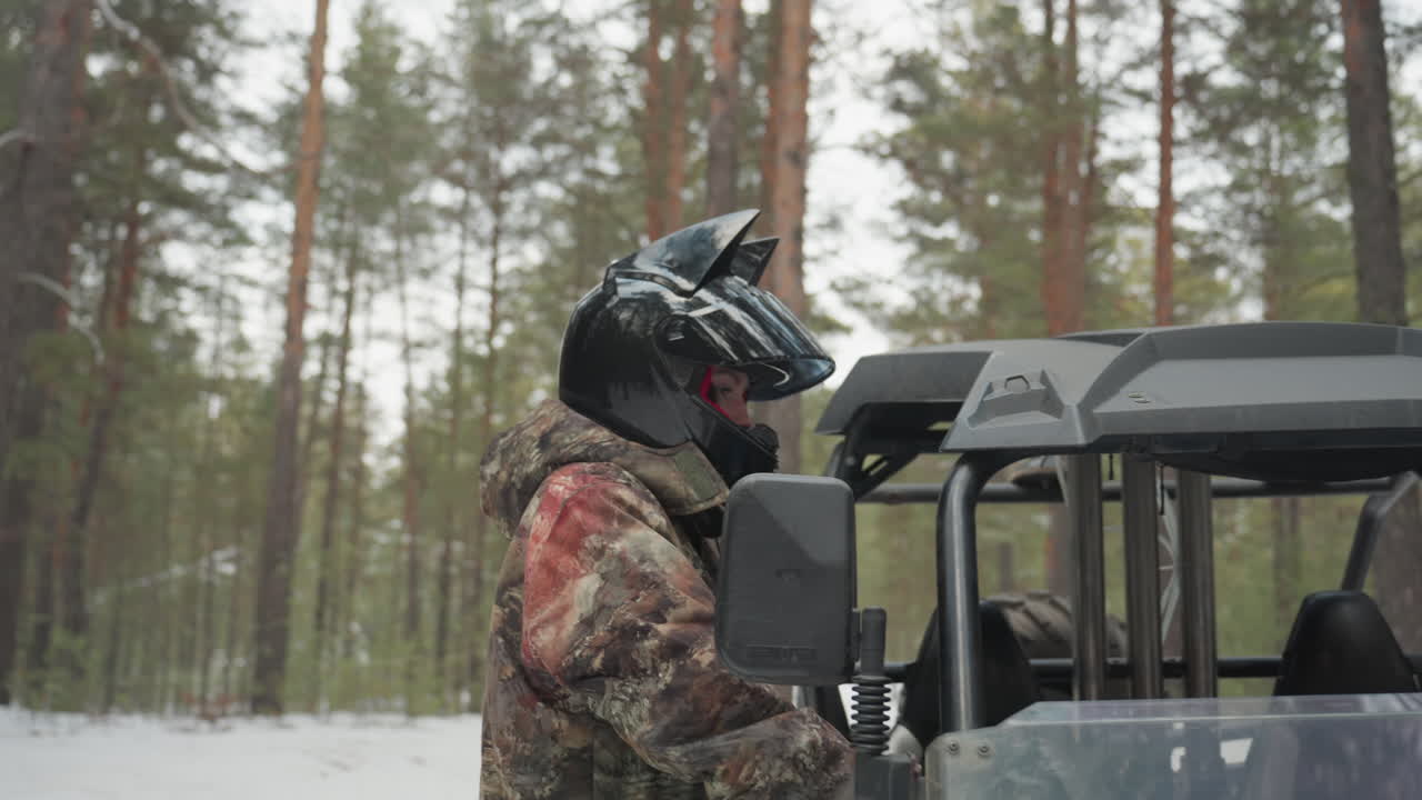 Piloti in una foresta innevata accanto a un UTV effettuano controlli pre-partenza e caricano l'equipaggiamento, la coppia con i caschi ispeziona le porte e il parabrezza, fissa il carico e regola le cinghie, veicolo utility blu con roll-bar.