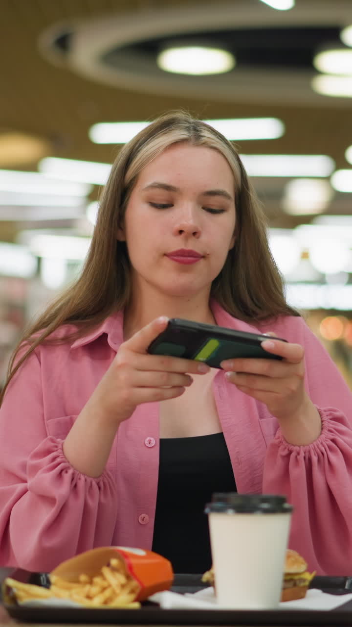 dama de vestido rosa se sienta en una mesa de madera en un restaurante bullicioso, tomando una foto de su comida, la comida incluye una hamburguesa, papas fritas y taza de café en una bandeja negra delante de ella, con un fondo borroso