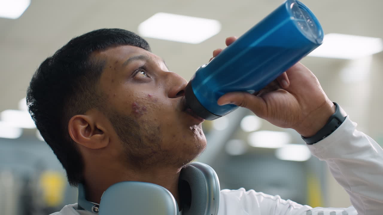 close up young man in white activewear drinking from blue water bottle after gym workout session with wireless headphones around neck and soft lighting in background