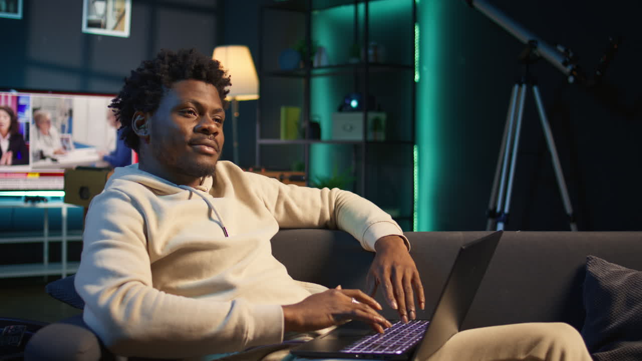 Young adult working remotely from his loft apartment on a laptop. Black freelancer focusing on tasks, blending productivity and relaxation while browsing the web from a cozy sofa. Camera B.