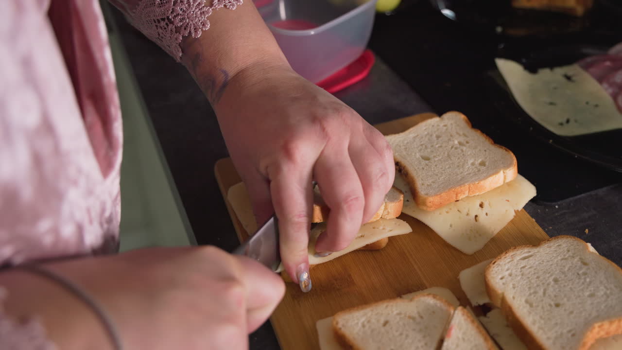 Caregiver in pink robe cuts cheese sandwiches with sharp knife on wooden board, preparing multiple meals on kitchen counter with visible lunch ingredients like deli slices, containers, and apples nearby