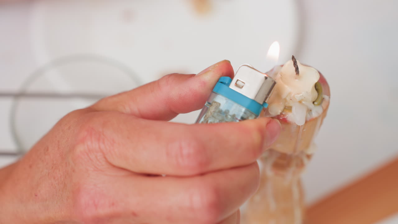 Close up of adult hand holding blue lighter igniting candle covered with melted wax drips, showing careful motion and warm flame during indoor moment