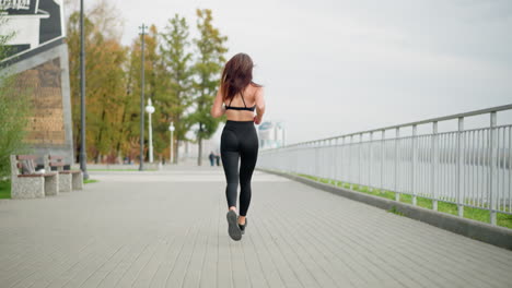 woman jogging in black athletic wear, running through park with iron fence, benches, and two blurred people in background, highlighting healthy lifestyle, fitness, and outdoor activity