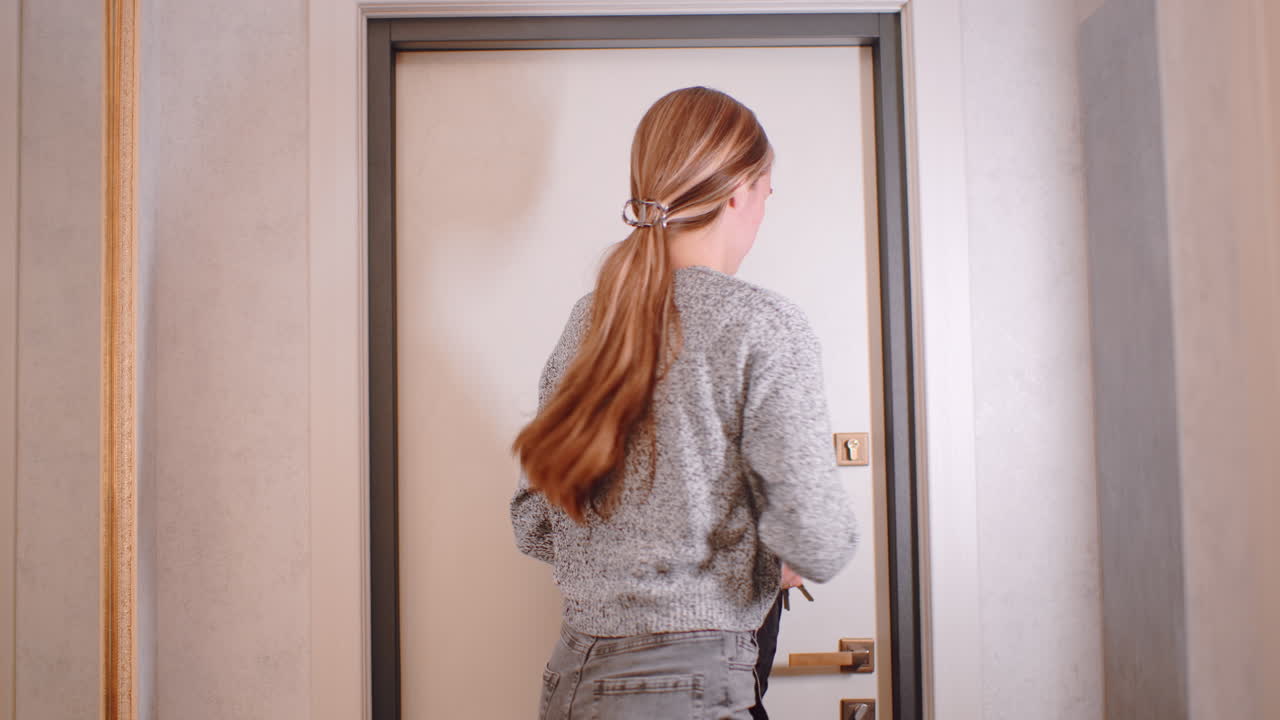 Young woman in hallway turns, grabs jacket, picks keys from hook, leave home, long hair in tie, casual gray sweater and jeans, front door behind, everyday independence
