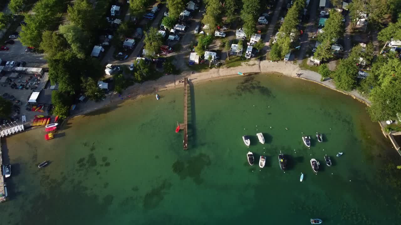 Aerial footage panning down over the top of a campsite beach and pier. Beautiful blue coloured water with a small number of boats at Lac Bleu at the bottom end of Lake Annecy in France