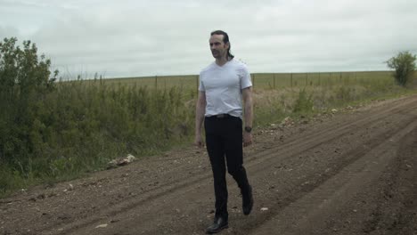 a man dressed in a white t-shirt and dark pants strolls along a dirt road surrounded by expansive green fields