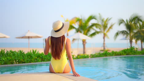 Backside-view-of-the-woman-sitting-on-edge-of-swimming-pool-at-exotic-hotel-in-Hawaii-near-the-beach-in-yellow-monokini-and-white-hat-and-hitting-water-with-her-legs,-beach-umbrellas-and-Palms