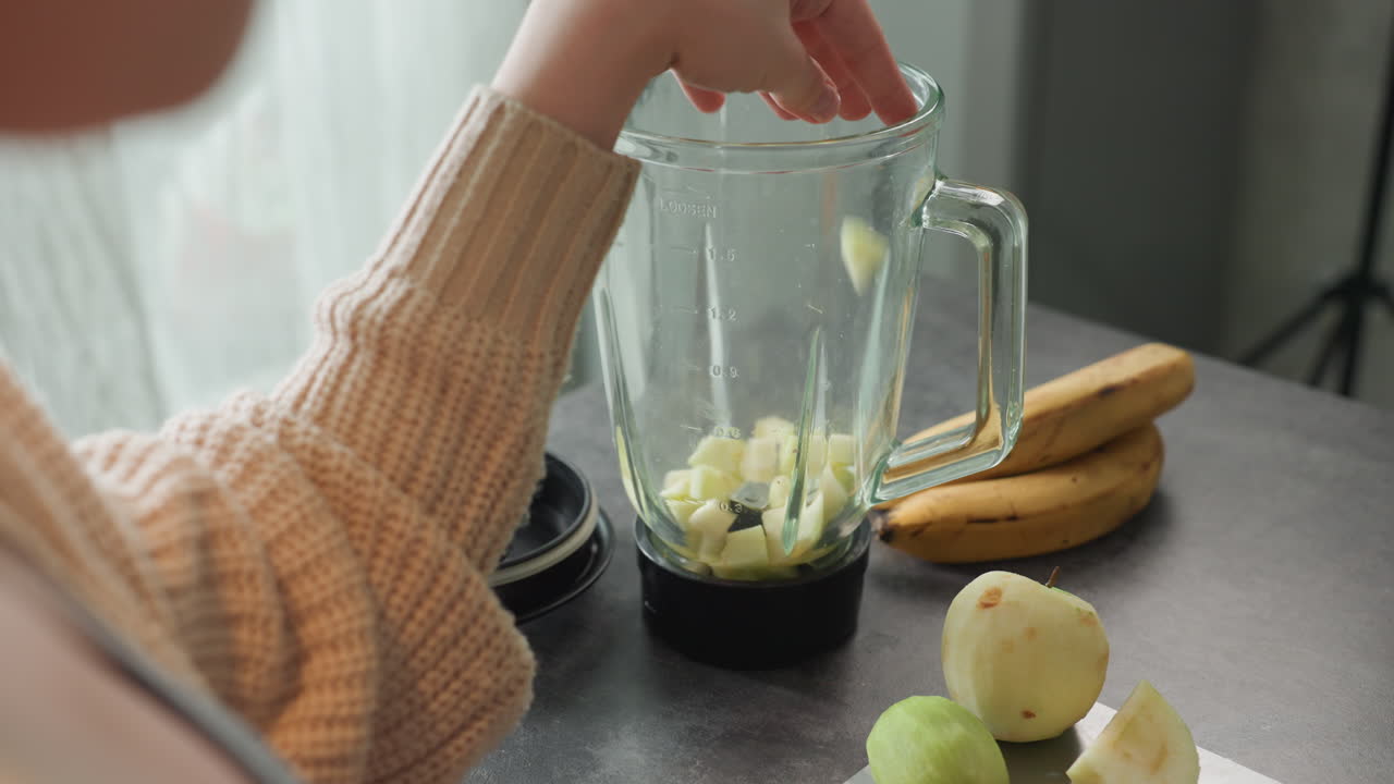Frullatore in primo piano con frutta, braccio di donna caucasica incinta che prepara un frullato in una cucina accogliente con luce soffusa del mattino, mele e banane sbucciate sul bancone, manica di maglione di lana, umore delicato e premuroso, concentrato.
