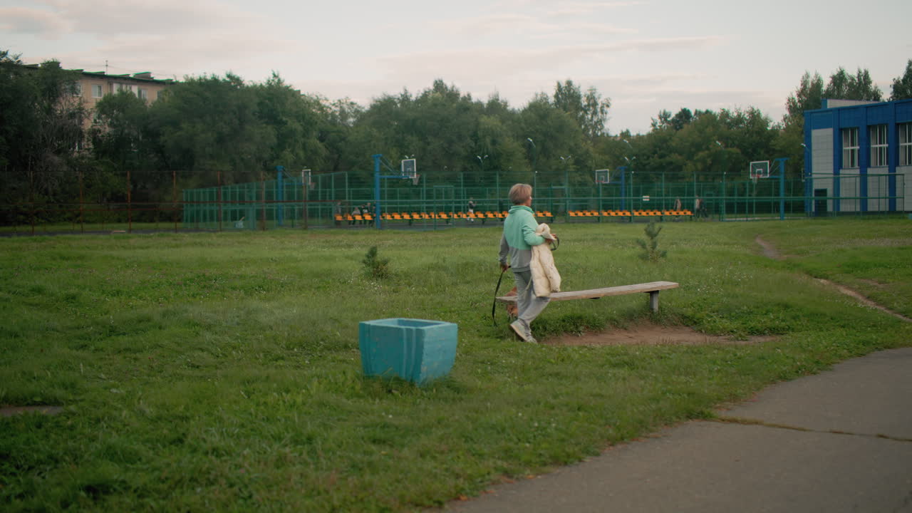 Canine specialist carrying jacket in hand walks with her playful puppy toward wooden bench on lush green field near outdoor basketball court surrounded by fence and trees on peaceful morning