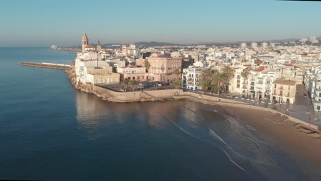 Drone-flies-along-side-touristy-coastal-city-lit-by-warm-morning-sun-as-gentle-sea-waves-quietly-roll-onto-the-shore