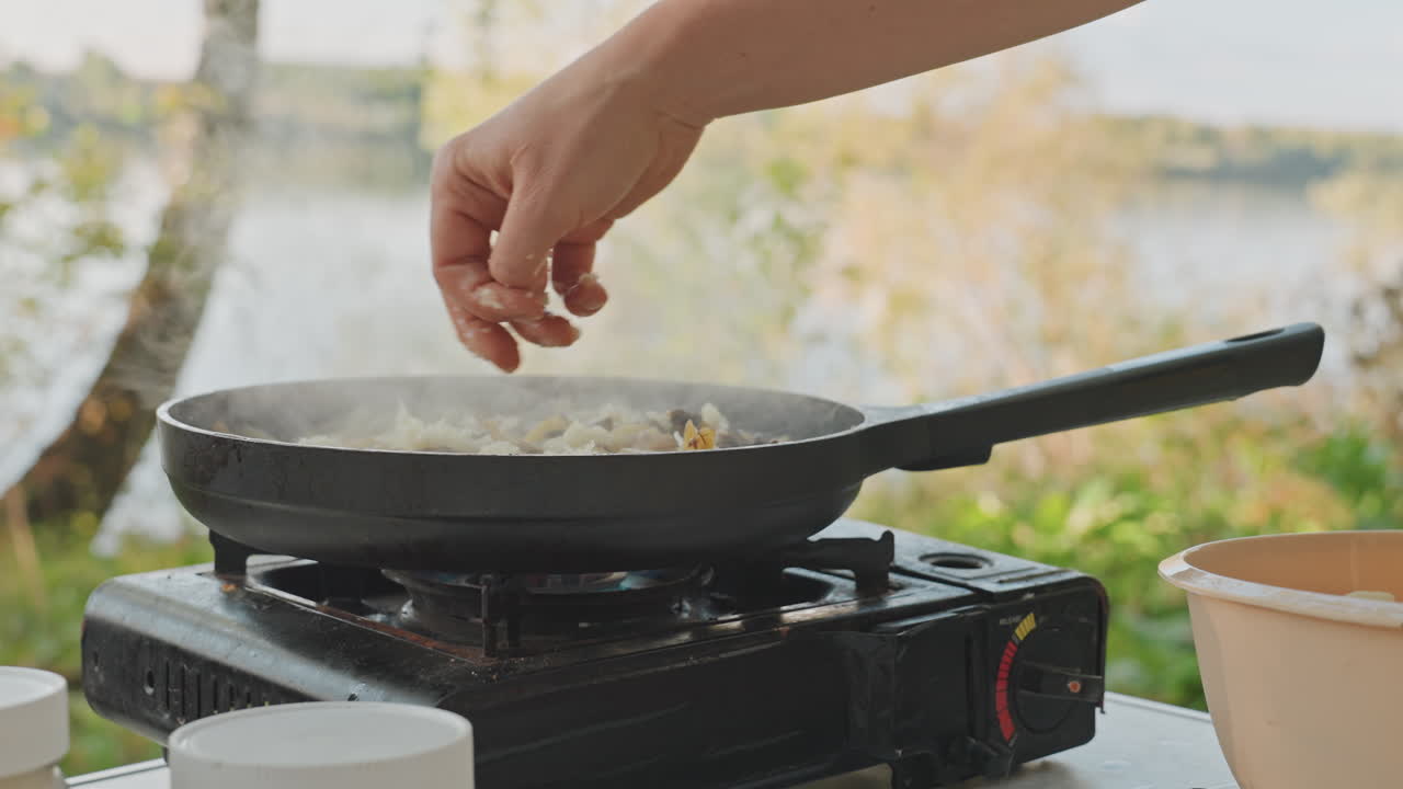 Campingpande krydret med salt og urter ved søbredden, delikat hånd drysser krydderi over sydende kartofler på bærbart blus, birketræer og vand i baggrunden, rolig morgenstemning.