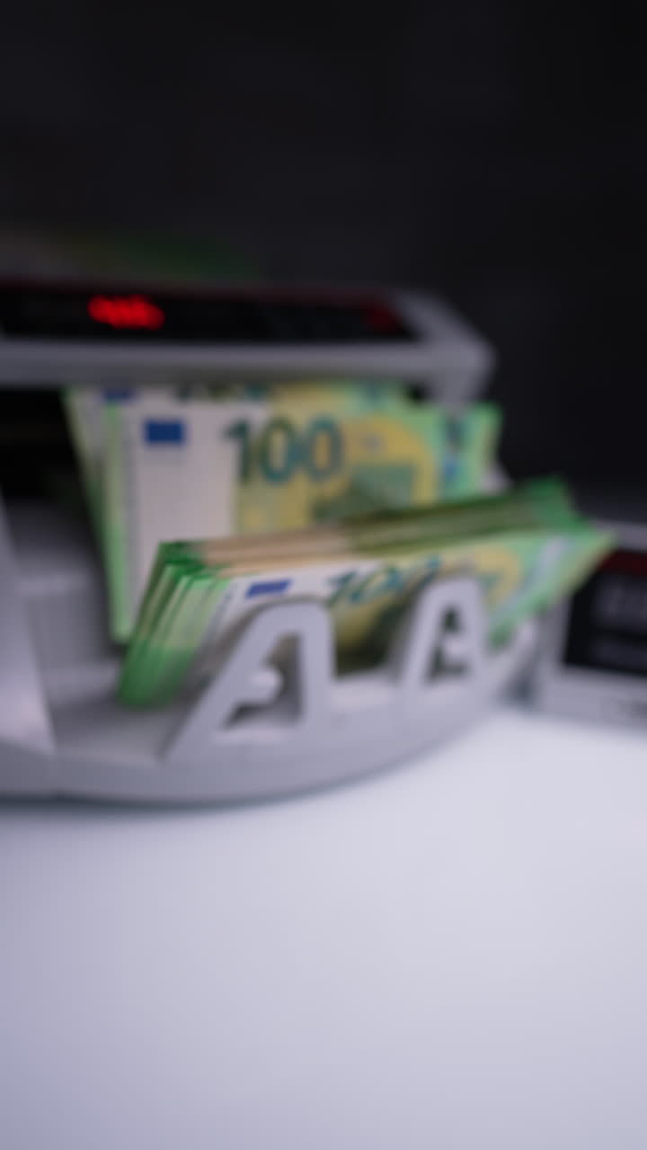 New green one hundred euro bills appear from the working cash counting machine. Male hand takes counted stack and puts on the desk. Vertical video.