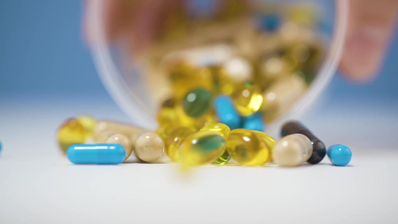 Molteplici pillole capsule multivitaminiche colorate che si riversano da una tazza su una superficie bianca, sfondo sfocato.