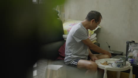 un viejo chino desayunando en el salón de un apartamento estrecho.