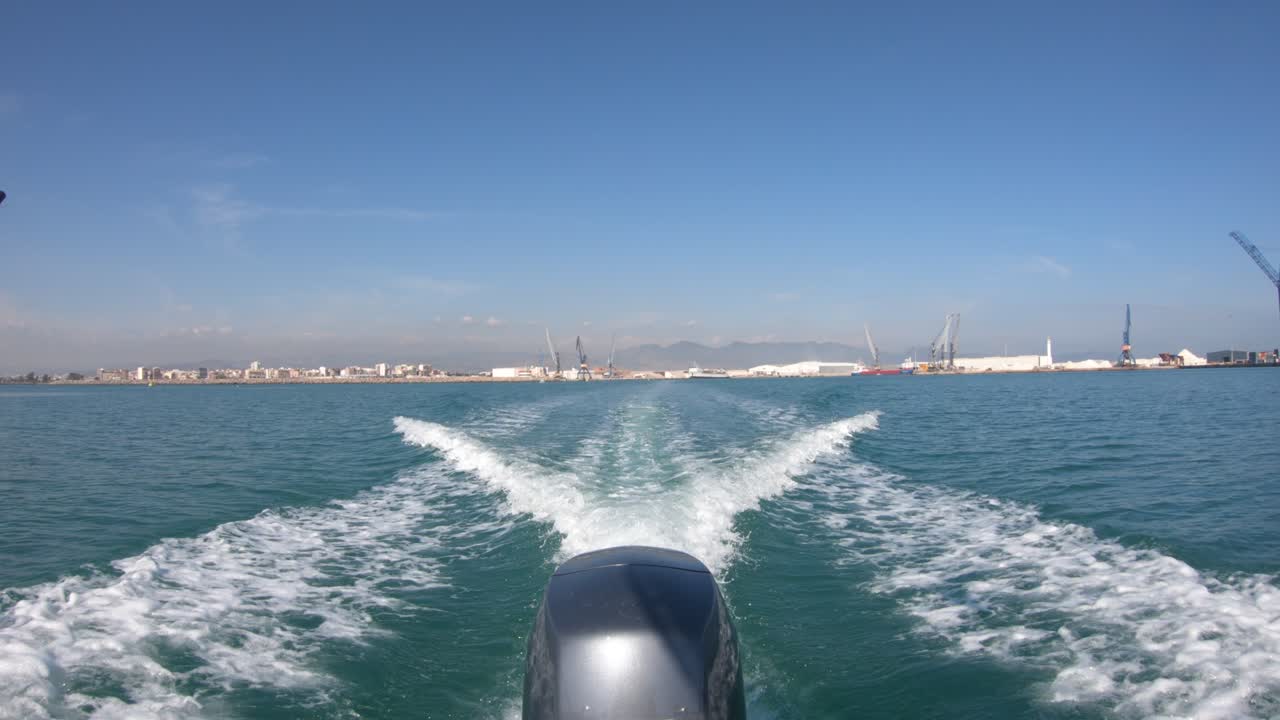 해안에서 멀리 이동하는 모터 보트의 선미, 크레인과 건물은 수평선에서 멀리 떨어져 있으며 엔진은 해수에서 웨이크를 그립니다.