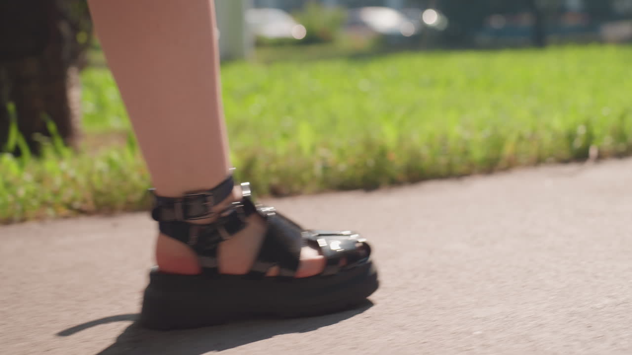 햇빛이 비치는 인도에서 검은색 샌들을 신고 걷는 십 대 백인 소녀의 발 클로즈업; 느리고 우울한 속도, 풀밭 가장자리, 가죽 스트랩과 페인트칠한 발가락이 보임, 도시 공원 배경, 얕은 피사계 심도