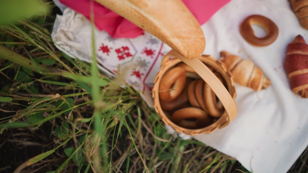 Close up of hand reaching into frame to pick bread from woven basket on white cloth with pink fabric and grass background. Basket contains bagels croissants various baked goods arranged