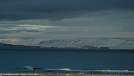 Leere-Arktische-Welle-Aus-Der-Ferne,-Die-Auf-Einem-Flachen-Riff-Landet,-Umgeben-Von-Schneebedeckten-Bergen