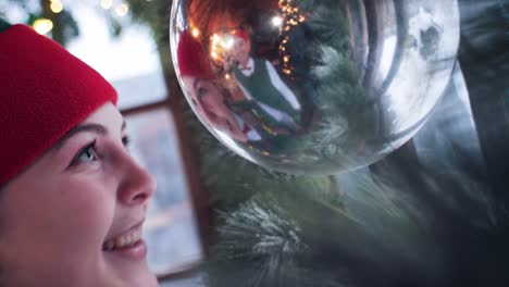 funny girl elf in a red hat looks in a new year s ball playing with its reflection. residence of santa claus