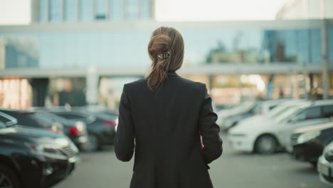 back view of professional woman walking with red folder and coffee cup in hand through city parking lot with blurred background of parked cars and glass residential building