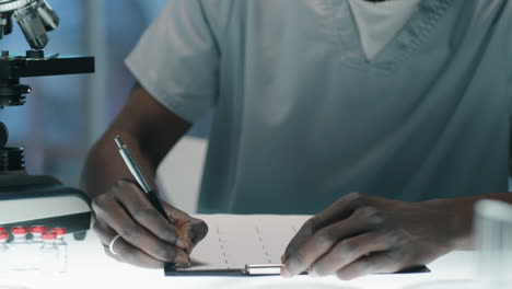 Trabajador-De-Laboratorio-Negro-Tomando-Notas-Y-Usando-Microscopio