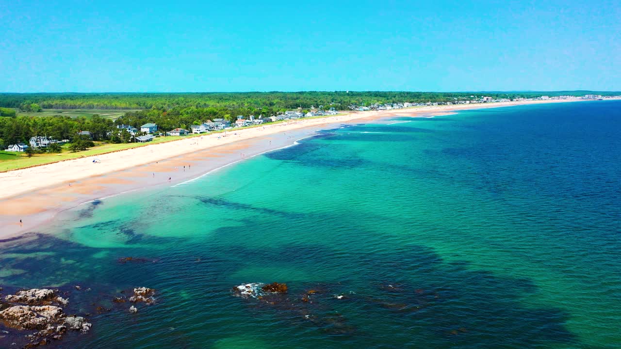 Drone view of Saco, Maine’s lively beaches. Crowds relax on warm sand as Atlantic waves crash near houses. East Coast travel vibe shows peak-season tourism, beautiful ocean, and coastal fun.
