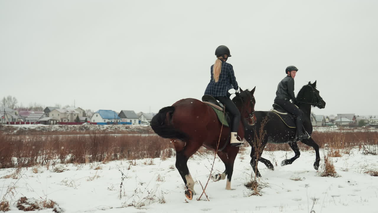 Due cavalieri che trottano insieme attraverso una pianura innevata, allenamento dinamico e collaborazione con l'allenatore nelle vicinanze, falcate ritmiche, caschi, selle, skyline del villaggio, vapore del respiro e lavoro di squadra invernale durante la cavalcata