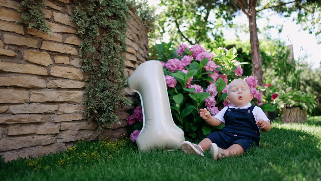 Cute blond Caucasian child sits on the grass alone. Little kid picks some grass and talks. Number one balloon is beside the baby.
