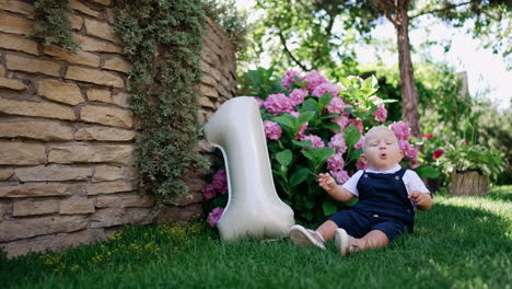 cute blond caucasian child sits on the grass alone. little kid picks some grass and talks. number one balloon is beside the baby.