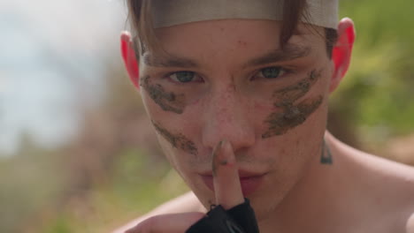 fierce young warrior with muddy streaks across face stares intensely while placing gloved finger over lips in silent command gesture, standing shirtless in sunlit outdoors, evoking strength