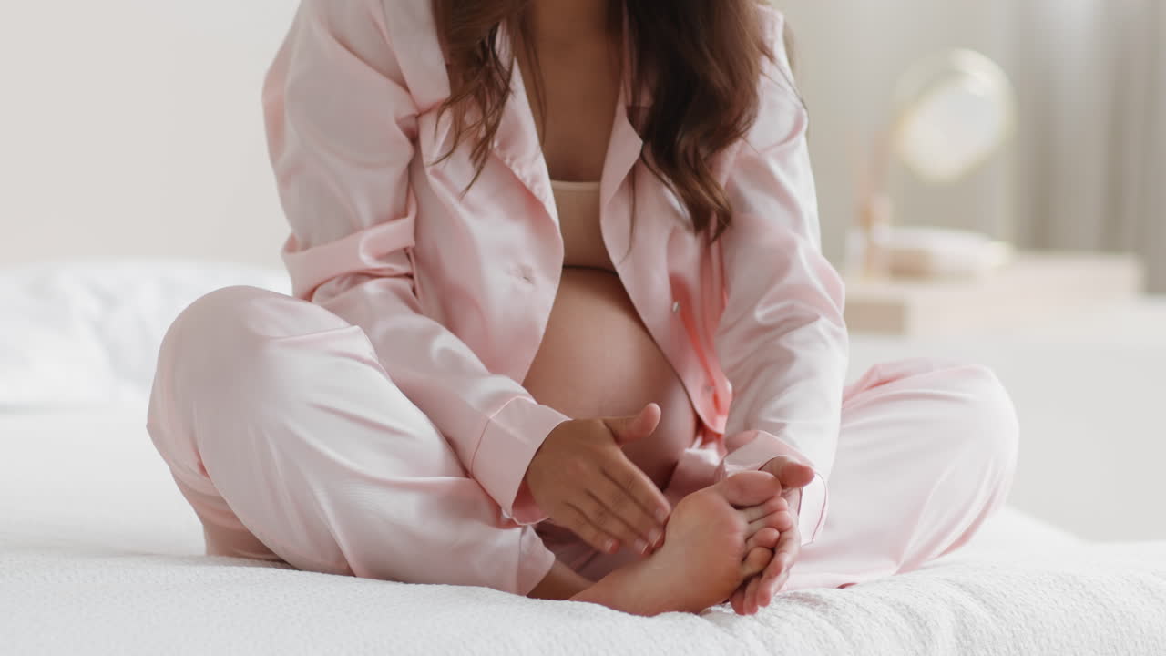 Pregnant Woman Massaging Her Feet in Satin Pajamas