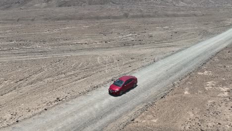 Toma-Panorámica-Aérea-De-Un-Vehículo-Rojo-En-Movimiento-En-El-Desierto-De-Perú-Frente-A-La-Ciudad-Santa-De-Caral-En-Una-Carretera-Polvorienta-Que-Domina-El-Paisaje-Seco-Con-Colinas-En-El-Fondo