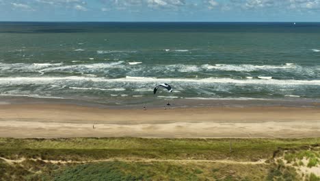 Toma-Panorámica-Aérea-Media-De-Un-Parapente-Volando-A-Lo-Largo-De-La-Playa-Sobre-Dunas-De-Arena-En-Un-Día-Soleado-Y-Ventoso-Con-Olas-De-Fondo