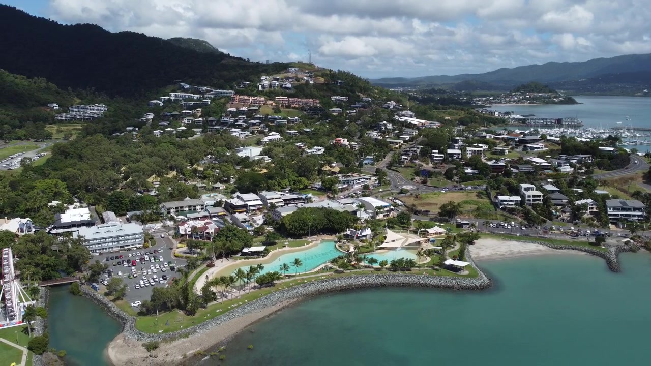 Vista aérea panorâmica destacando a famosa lagoa pública de Airlie Beach, o parque à beira-mar e a paisagem urbana costeira com colinas tropicais ao fundo