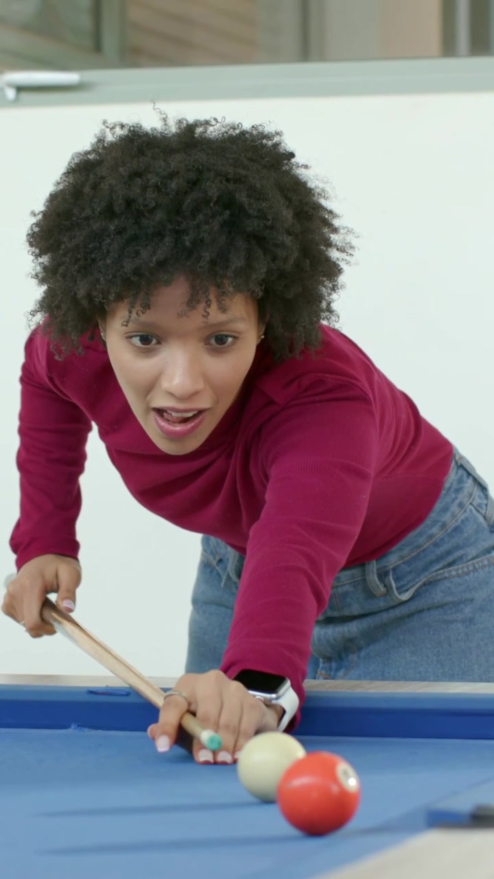 Vertical video: Leaning woman aiming and striking cue ball at blue pool table for fun, burgundy top