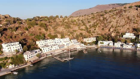 drone view in greece flying over blue sea in loutro small white house town and small boats next to a hill on sunset light