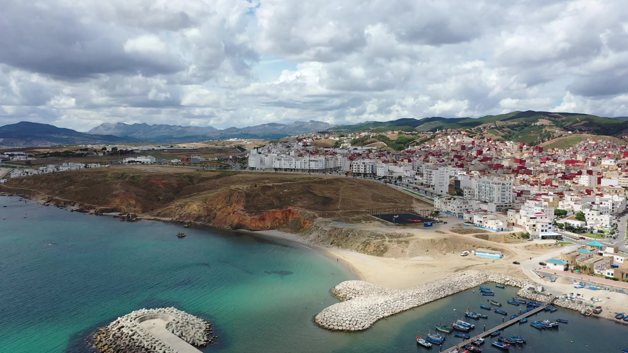 Ampia vista aerea del porto peschereccio di M'diq, frangiflutti, acqua turchese del Mediterraneo e quartieri costieri sotto i Monti del Rif nel Marocco settentrionale, Nord Africa