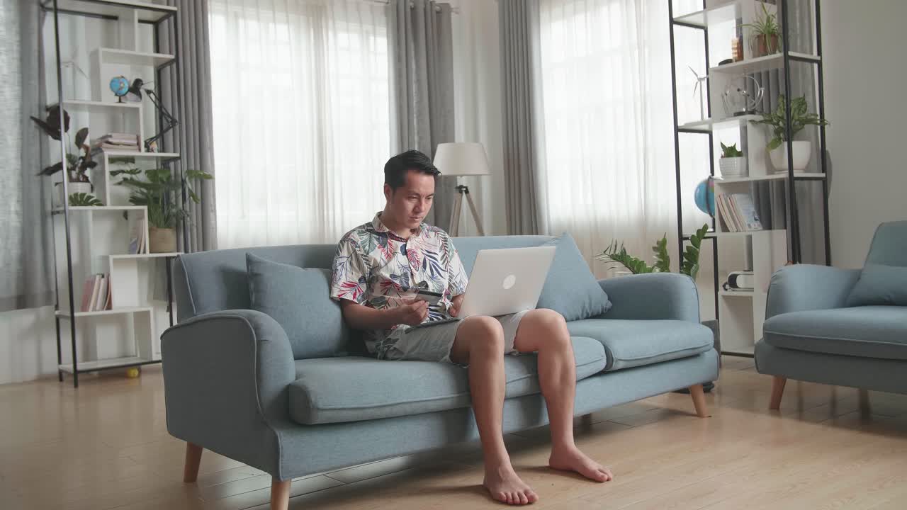 Asian Man Sitting On Couch In Living Room, Hand Holding Credit Card And Using Laptop Computer Shopping Online