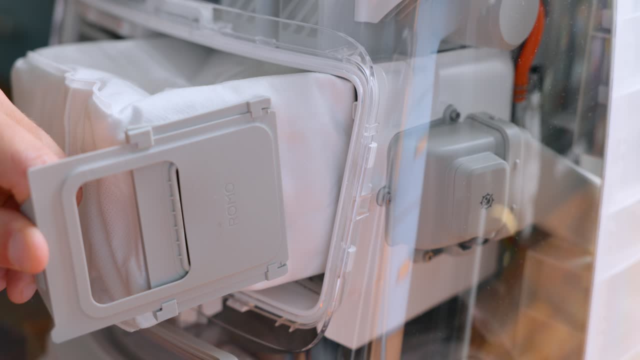 Close-up of hands opening a robot vacuum dock and removing the dust filter bag from the compartment