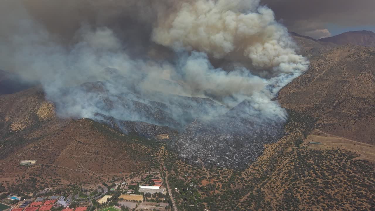 Vista aerea cinematografica con drone di un incendio che si propaga lungo il fianco di una montagna arida verso edifici e infrastrutture nella zona di confine urbano di Santiago, Cile, video 4K