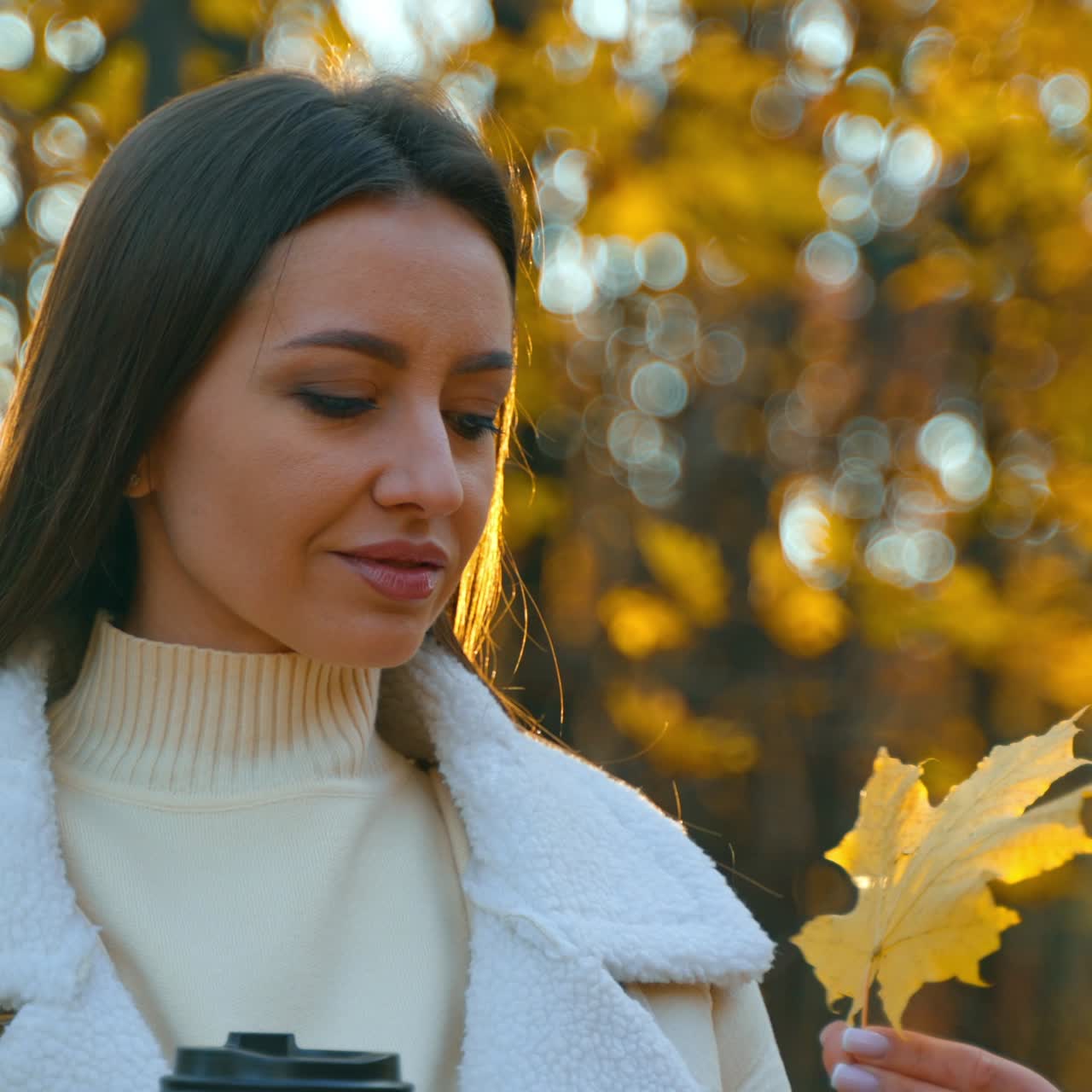 Beautiful lady with a paper cup in her hand covering half of her face behind a yellow maple tree leaf. Woman looks at the leaf turning it and raising to the sun. Close up. Blurred autumn backdrop
