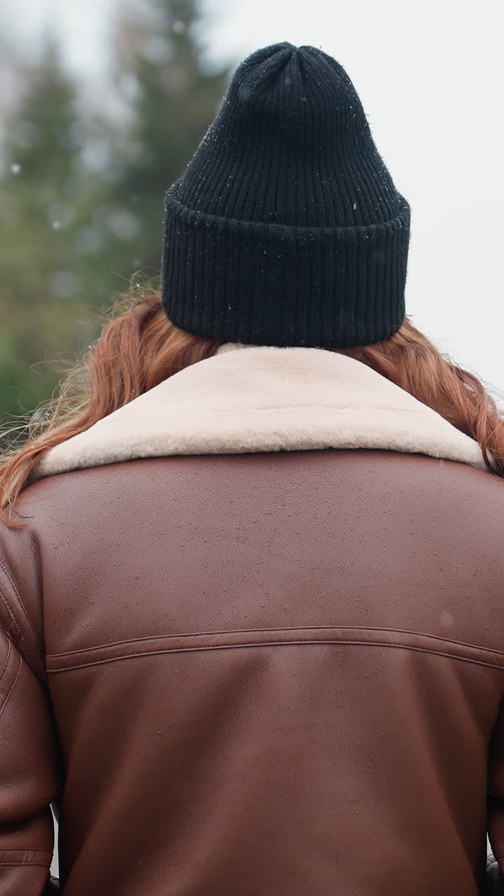 Back view of young woman in black cap and brown shearling jacket walking slowly with hands in pockets on wide stone pathway through city park during calm snowy day