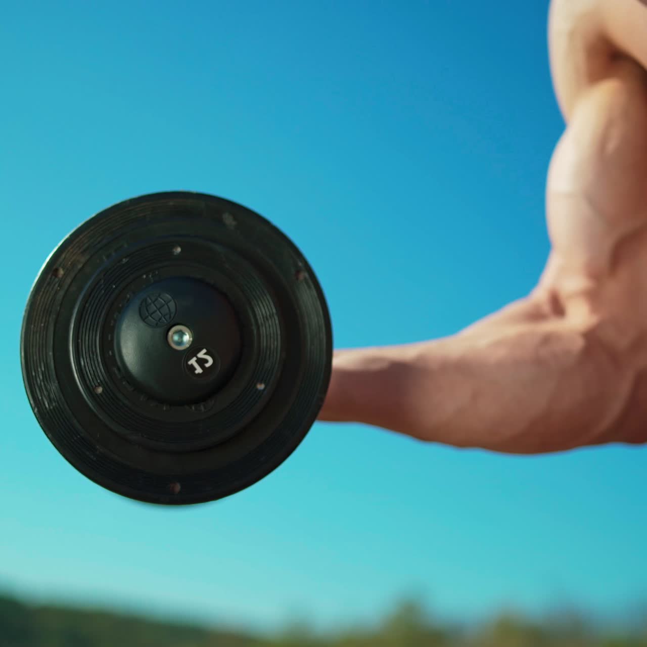 Masculine arm with metal dumbbell. Athletic man without shirt with muscular body lifting heavy weights by hand on blue sky background. Close-up.