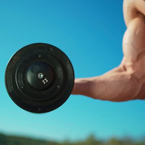 masculine arm with metal dumbbell. athletic man without shirt with muscular body lifting heavy weights by hand on blue sky background. close-up.