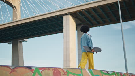 relaxed teenager walking skateboard under city bridge.  skater relaxing