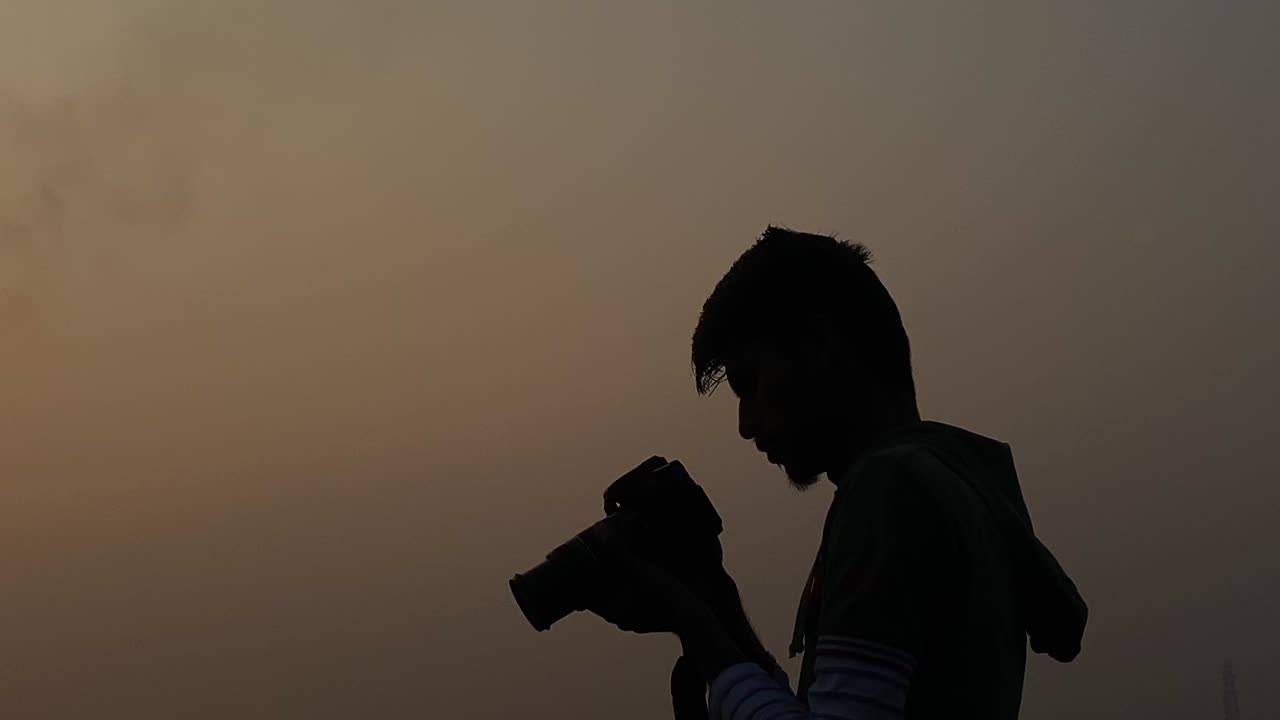 夕暮れの曇り空の下、シルエットで静止した横顔の男性がカメラを構えて撮影。