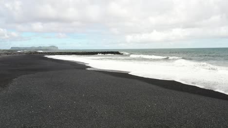 Playa-De-Arena-Negra-Con-Olas-Y-Rompeolas-En-Vik,-Islandia-Con-Video-De-Drones-Bajo-Y-Avanzando