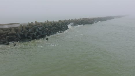 Aerial-establishing-view-of-Port-of-Liepaja-concrete-pier,-Baltic-sea-coastline-,-foggy-day-with-dense-mist,-moody-feeling,-big-storm-waves-splashing,-wide-ascending-drone-shot-moving-forward