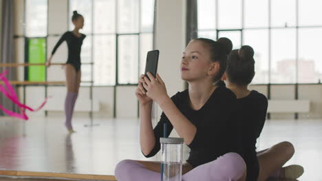 Gymnastisches-Blondes-Mädchen,-Das-Auf-Dem-Boden-Sitzt-Und-Ein-Foto-Von-Ihrer-Freundin-Macht,-Die-Tanzt-Und-Eine-Band-Hält-1